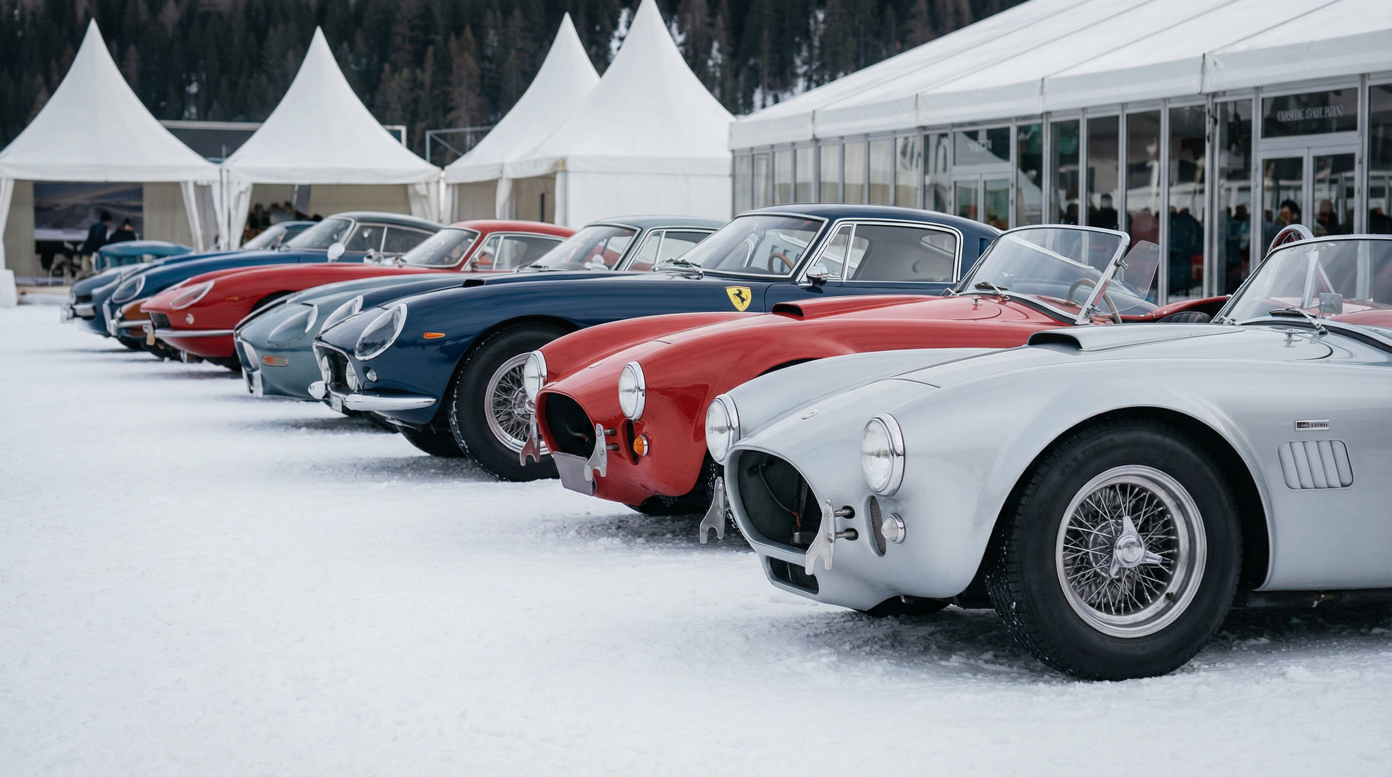 Vintage cars lined up on a snowy surface with tents and buildings in the background.