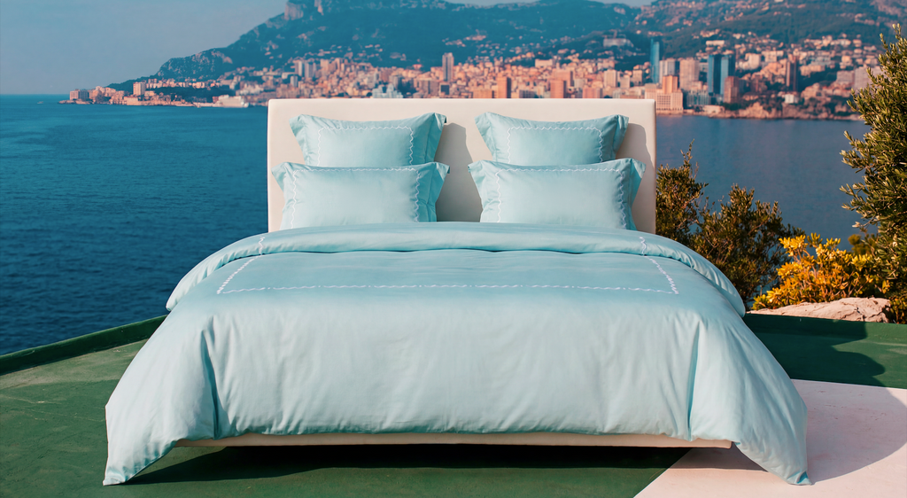 Bed with light blue bedding overlooking a city by the sea