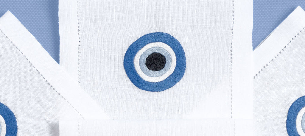 A set of white hand embroidered drink placemats with a blue Greek eye design on a blue background, displayed folded.
