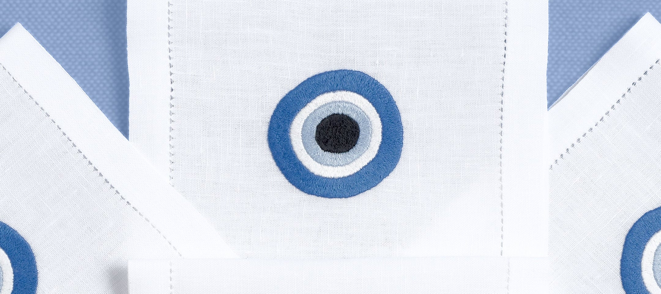 A set of white hand embroidered drink placemats with a blue Greek eye design on a blue background, displayed folded.