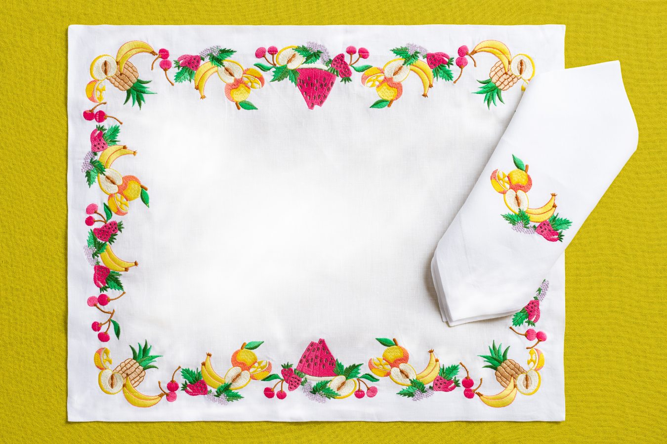 A rectangular placemat with a 'Tutti Frutti' theme, featuring colorful fruit designs on a white background, displayed on a yellow background with a part of the placemat showing a white napkin.