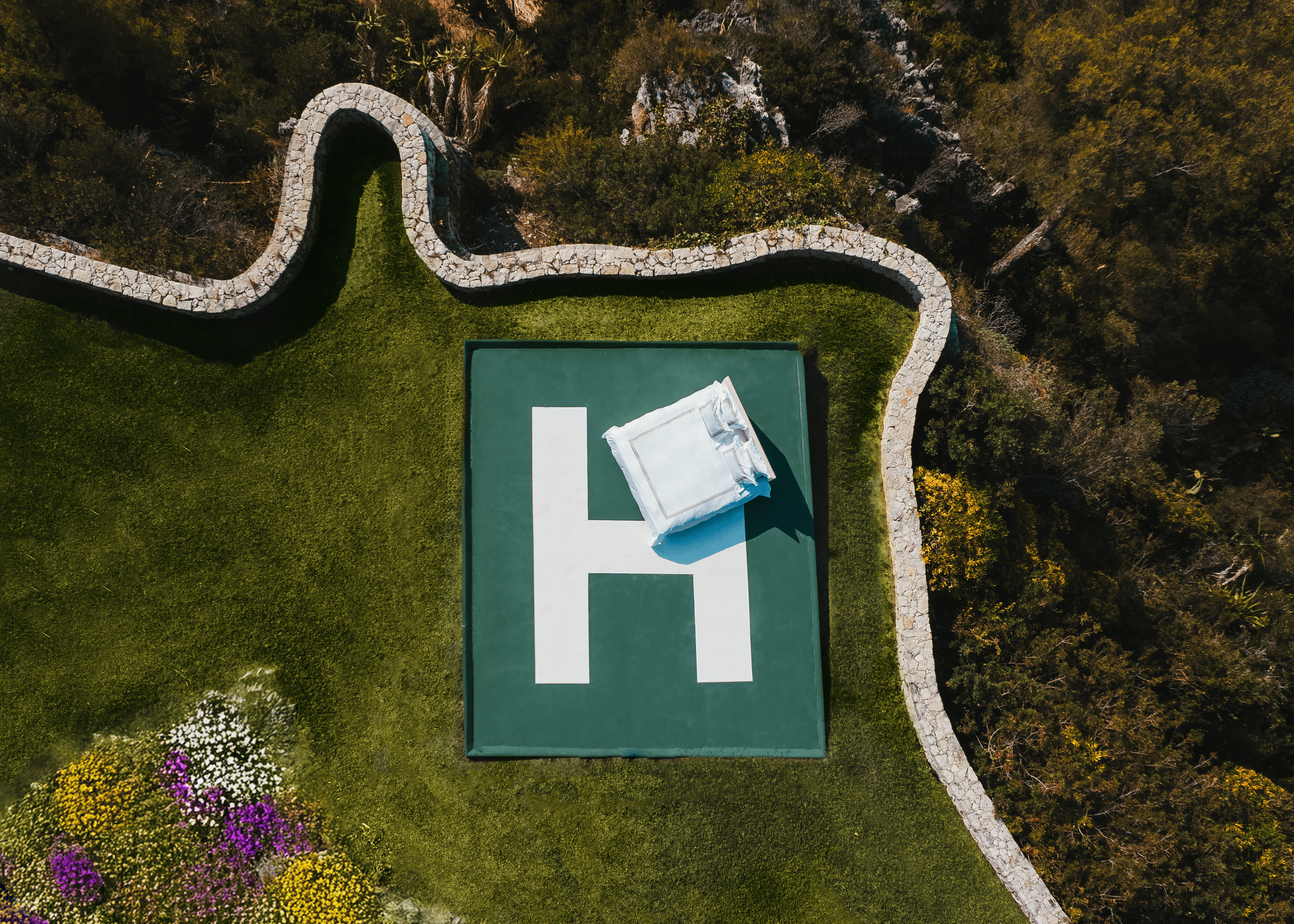 Aerial view of a green square with a white 'H' on a grassy area surrounded by trees and flowers.