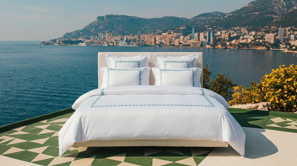 Bed with white bedding and decorative pillows by a scenic waterfront with mountains and cityscape.