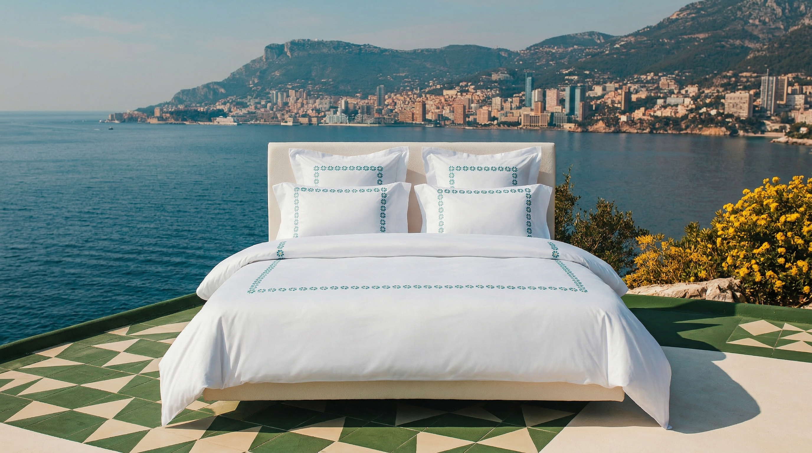 Bed with white bedding and decorative pillows by a scenic waterfront with mountains and cityscape.